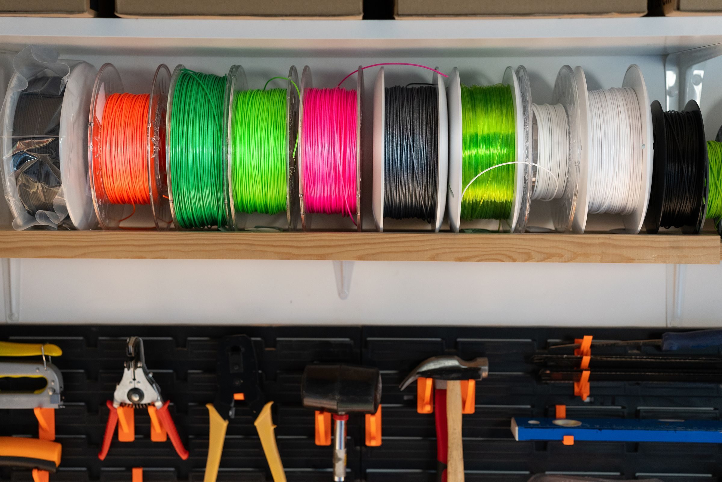 Atölye rafında yan yana dizilmiş turuncu, yeşil, pembe, siyah ve beyaz 3D yazıcı filament makaraları; altta duvar panosuna asılı el aletleri.