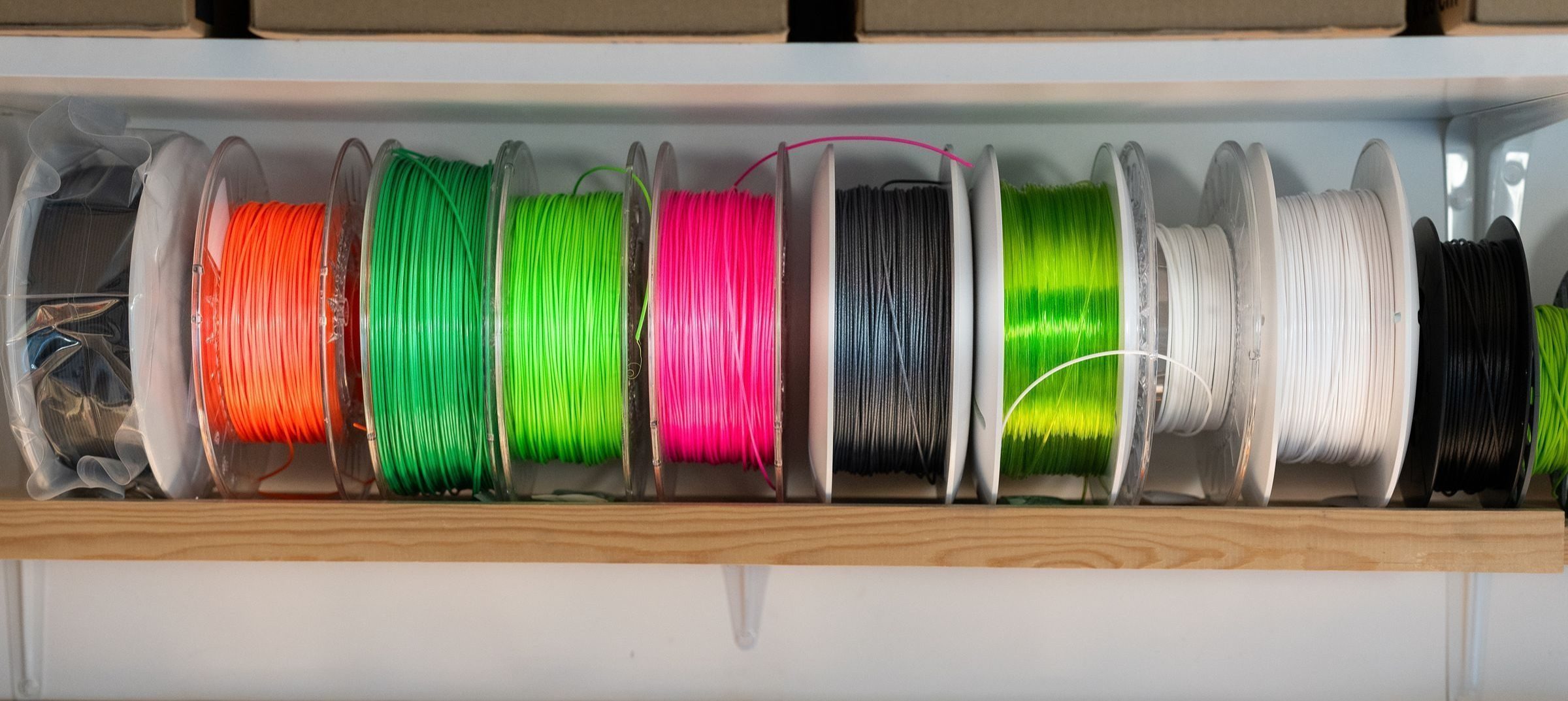 Atölye rafında yan yana dizilmiş turuncu, yeşil, pembe, siyah ve beyaz 3D yazıcı filament makaraları; altta duvar panosuna asılı el aletleri.