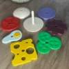 Montessori hamburger sıralama oyunu parçaları; domates, yumurta, peynir, salatalık, köfte gibi renkli 3D baskı katmanlar ve dikme çubuğu masa üzerinde dağınık şekilde sergileniyor.