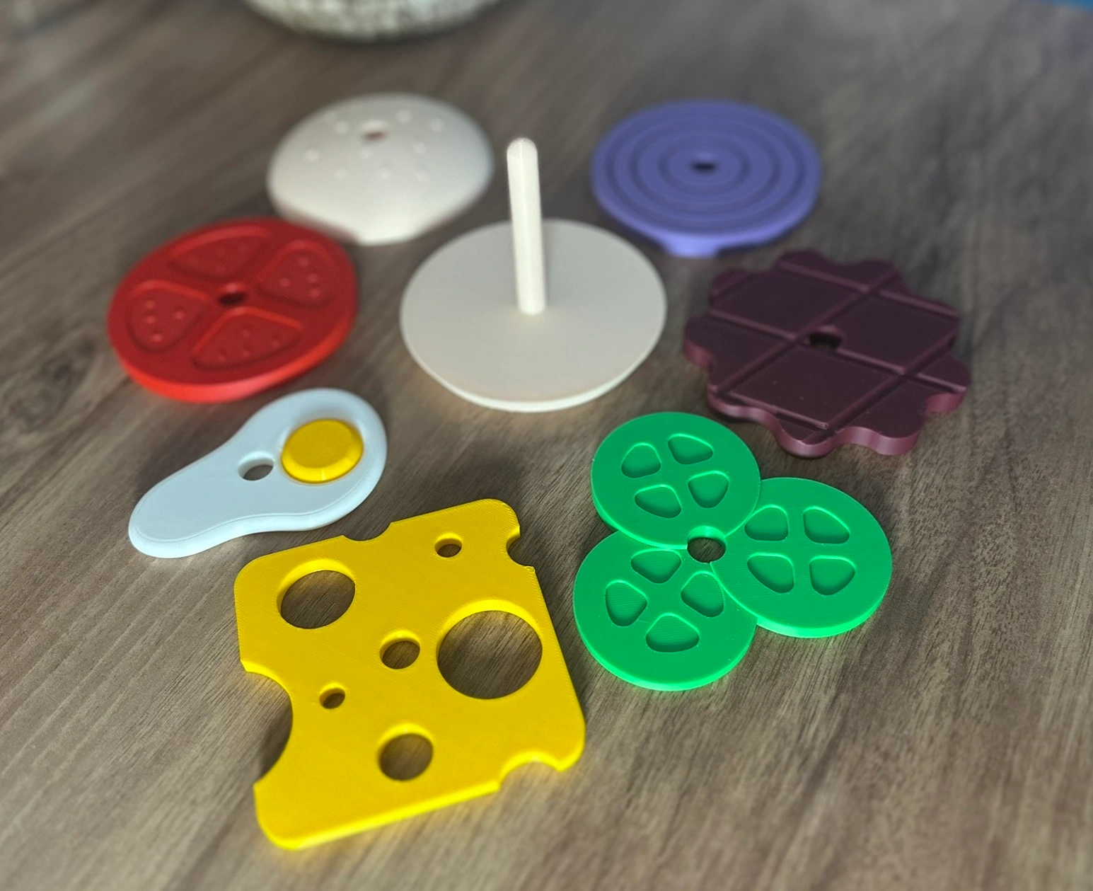 Montessori hamburger sıralama oyunu parçaları; domates, yumurta, peynir, salatalık, köfte gibi renkli 3D baskı katmanlar ve dikme çubuğu masa üzerinde dağınık şekilde sergileniyor.